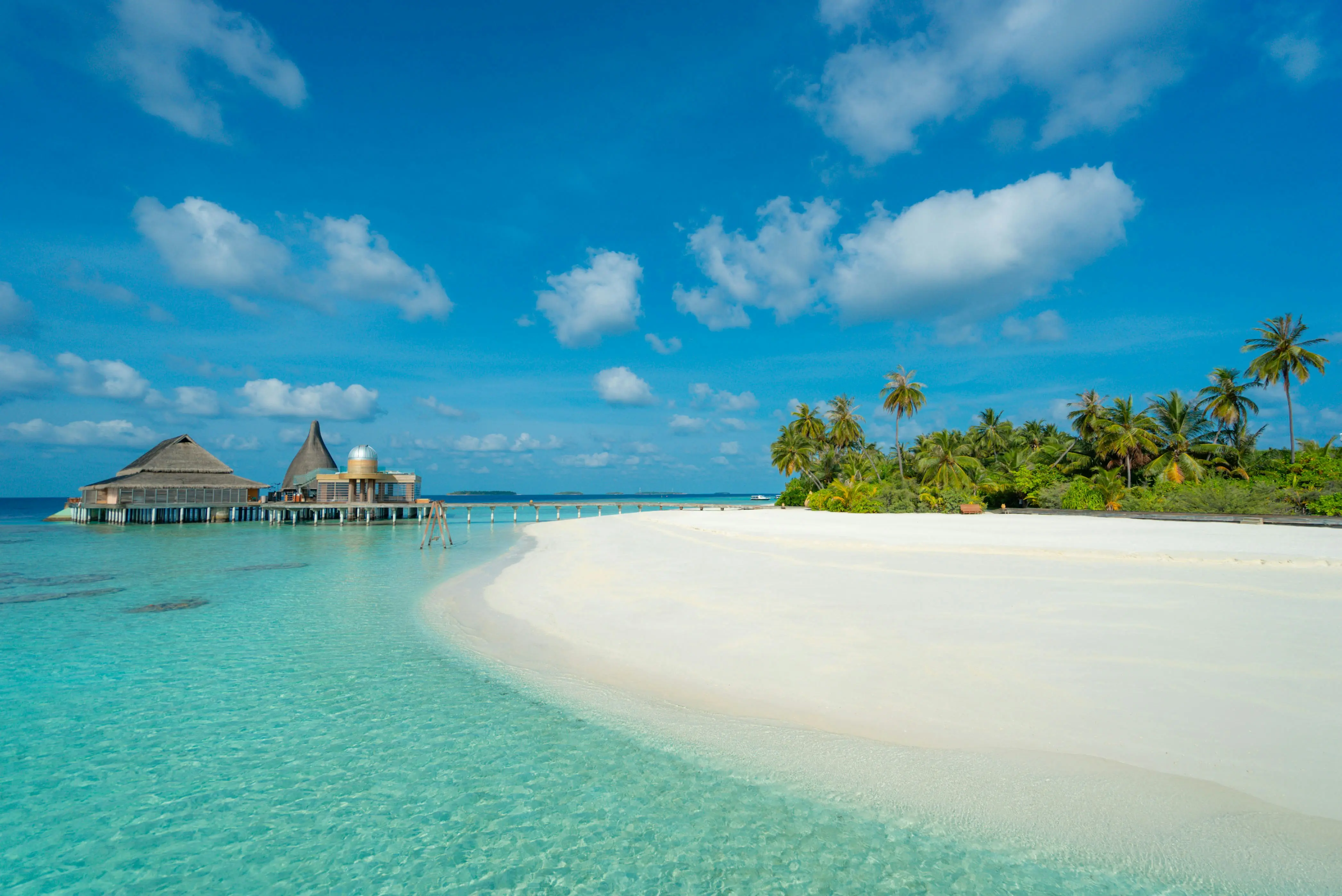 White sand beach, palm trees and an overwater building beside a turquoise lagoon in the Maldives